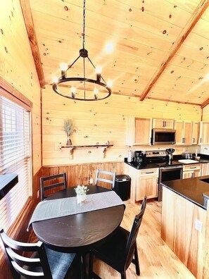 A wooden interior with a dining table, chairs, a chandelier, and kitchen appliances.