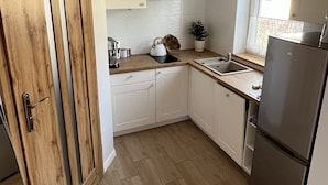 Eine kleine Küche mit weißen Schränken, einer Holztür, einem Edelstahlkühlschrank und einem Fenster mit Spitzgittervorhängen.