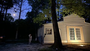 Une petite cabane en bois, d’un seul étage, avec un toit à pignon, entourée d’arbres et posée sur un sol en terre battue.