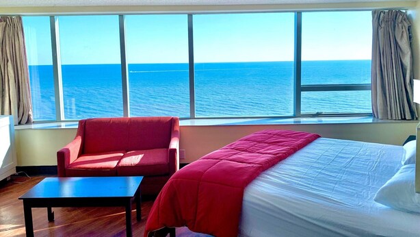 A hotel room with a large window offering a sea view, a bed, a red armchair, and a small table.