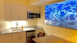 A compact kitchen with white cabinets, a stainless steel refrigerator, a microwave, and a sink. There's a small table with a chair, and a large TV mounted on the wall displaying a mountainous landscape.