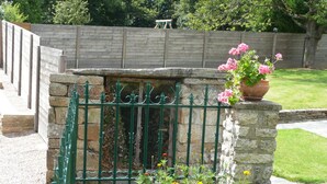 A stone wall with a green metal gate, a flower pot with pink flowers, and various plants in the foreground. - Country house with indoor pool, near Rochefort en terre; 2 stars. (Saint-Gravé)