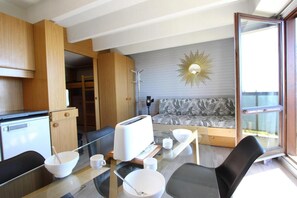 Un espace de vie compact comprenant un coin cuisine, un coin repas avec des chaises et une table en verre, ainsi qu’un canapé à motifs tropicaux.