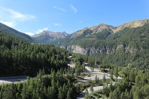 A mountainous landscape with a road leading through a forested valley, with buildings visible on the right side.