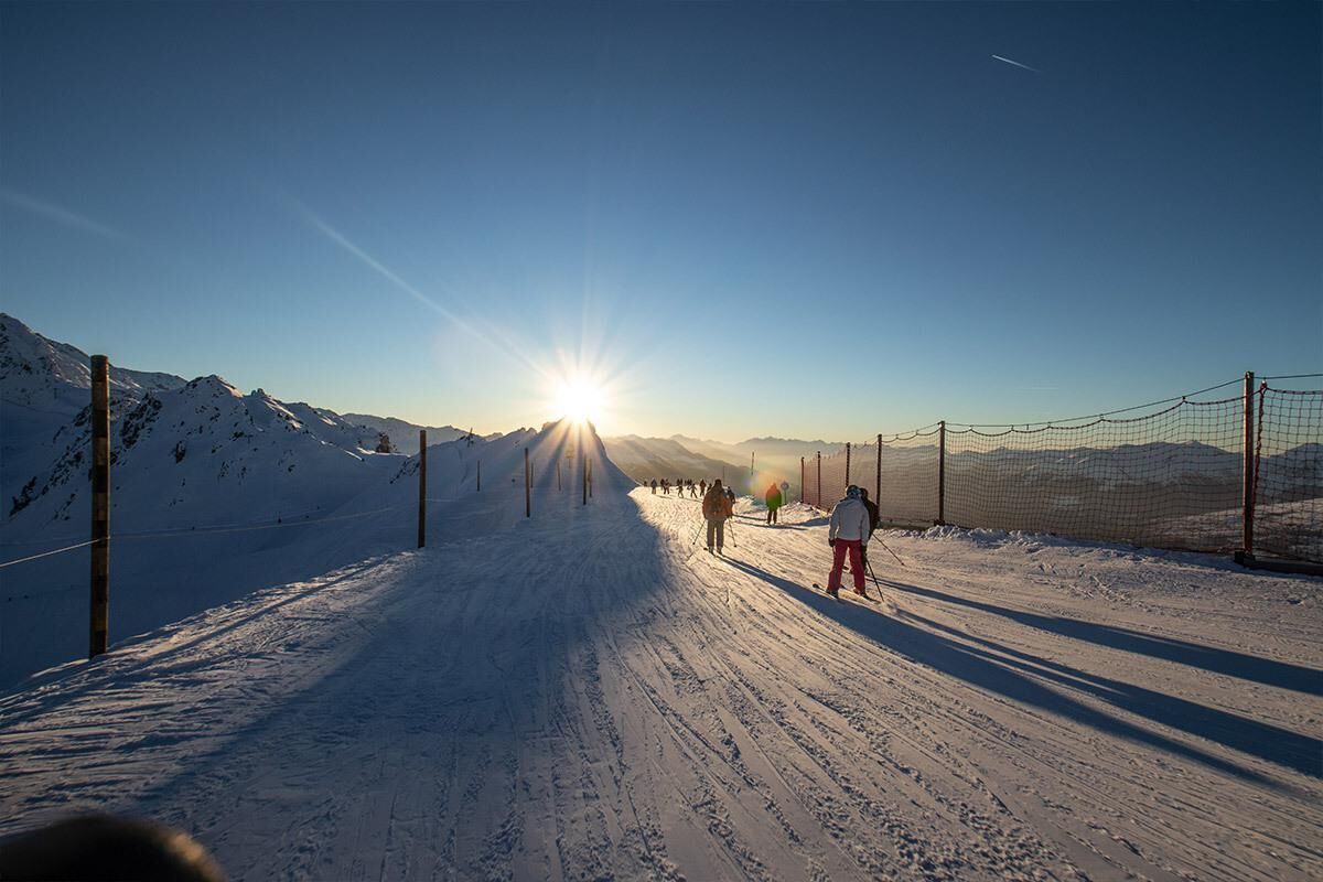 Uma estrada montanhosa coberta de neve, com céu claro e o sol brilhando.