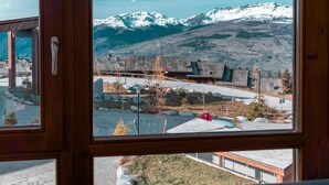 A room with a large window offering a view of snow-capped mountains and a building with a stone facade. - Ski-in/ski-out & foot of the slopes, 2nd floor, view open, fitness, swimming pool, balcony (Les Arcs)