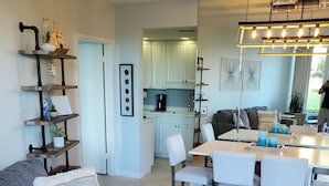 Een moderne woonkamer met een grijze bank, een salontafel met decoratieve objecten, een eethoek met een witte tafel en stoelen, en een keuken met witte kastjes en een koffieapparaat.