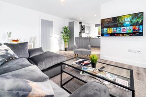 Una sala de estar moderna con un sofá gris, una mesa de centro de vidrio con revistas, un televisor que muestra opciones de películas y una cocina al fondo.