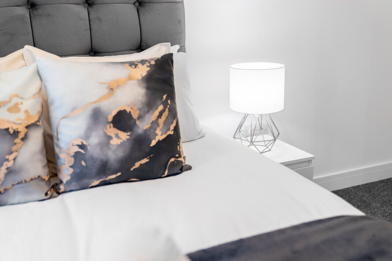 A neatly made bed with a grey upholstered headboard, a white bedside table with a geometric lamp, and two patterned pillows.