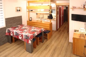 A compact room with a bunk bed, a small kitchenette, a dining table with a patterned tablecloth, and a television on a wooden stand.