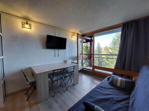 A room with a bed, a desk with two chairs, a TV mounted on the wall, and a balcony with a view of trees.