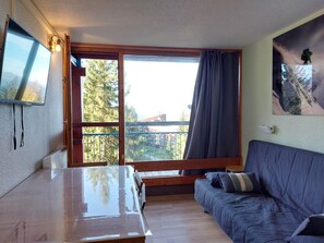 A room with a wooden table, a blue sofa, a TV mounted on the wall, and a large window with a view of trees and buildings.