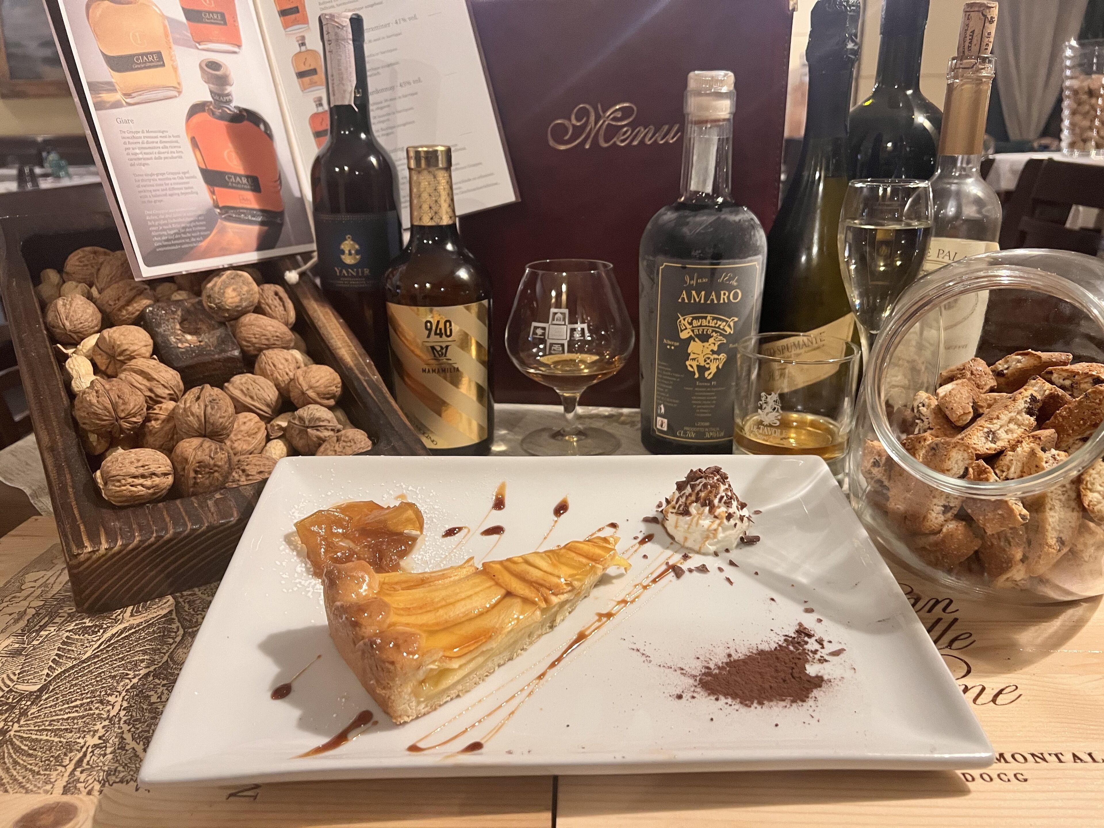 Um prato de sobremesa com uma fatia de torta, uma garrafa de vinho e um pote de nozes sobre uma mesa.