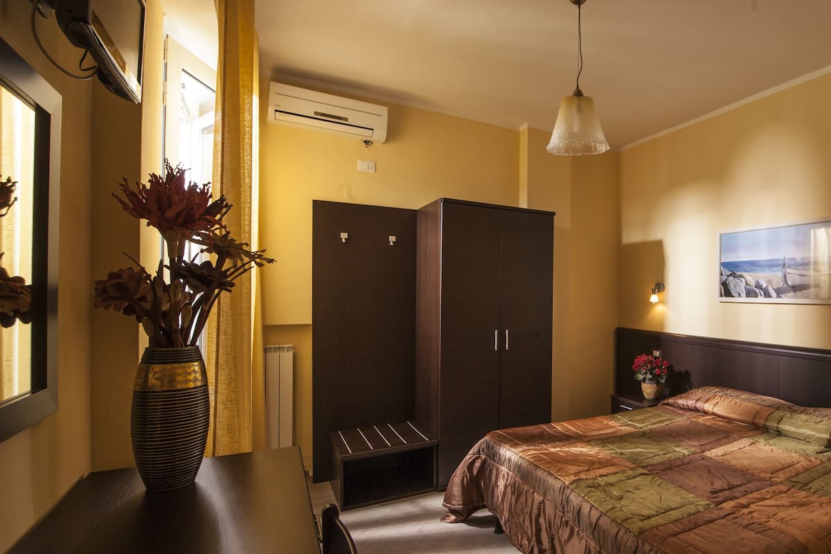 A hotel room with a bed, a nightstand, a dresser, a vase with flowers, and a wall-mounted picture.
