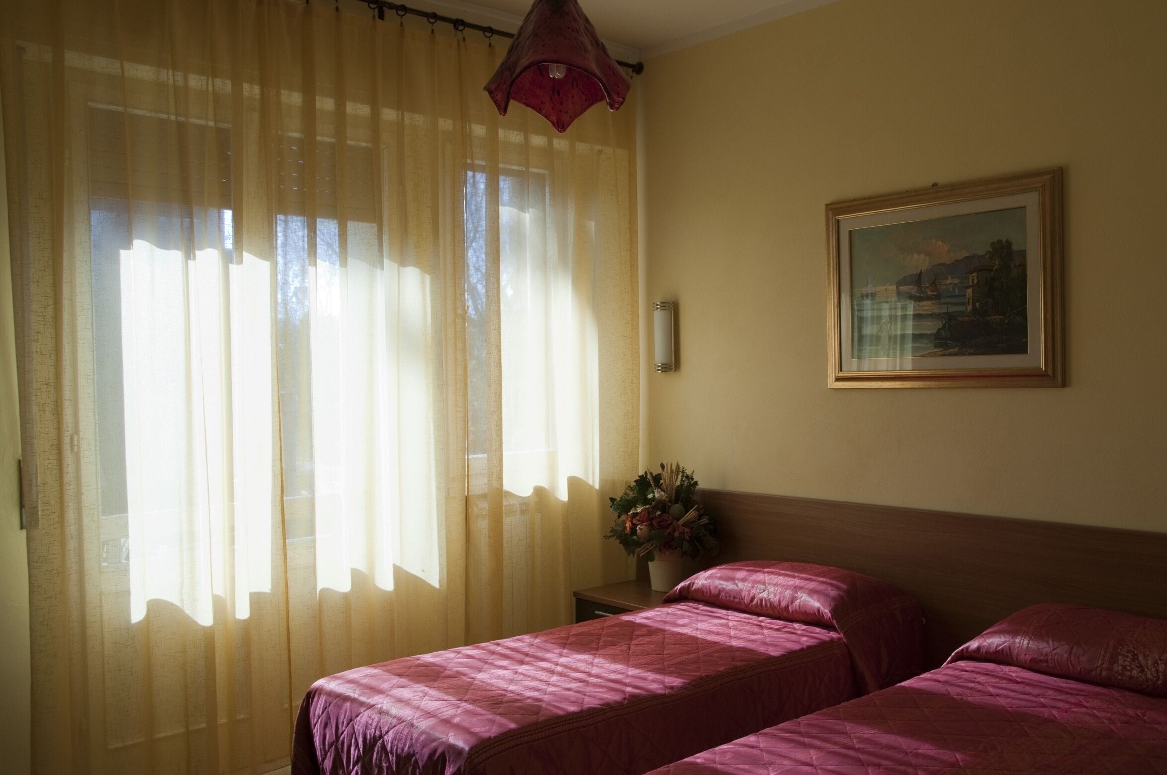A hotel room with two beds, a window with sheer curtains, a bedside table with a vase of flowers, and a framed picture on the wall.