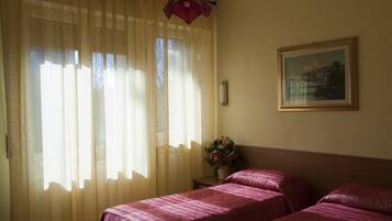 A hotel room with two beds, a window with sheer curtains, a bedside table with a vase of flowers, and a framed picture on the wall.