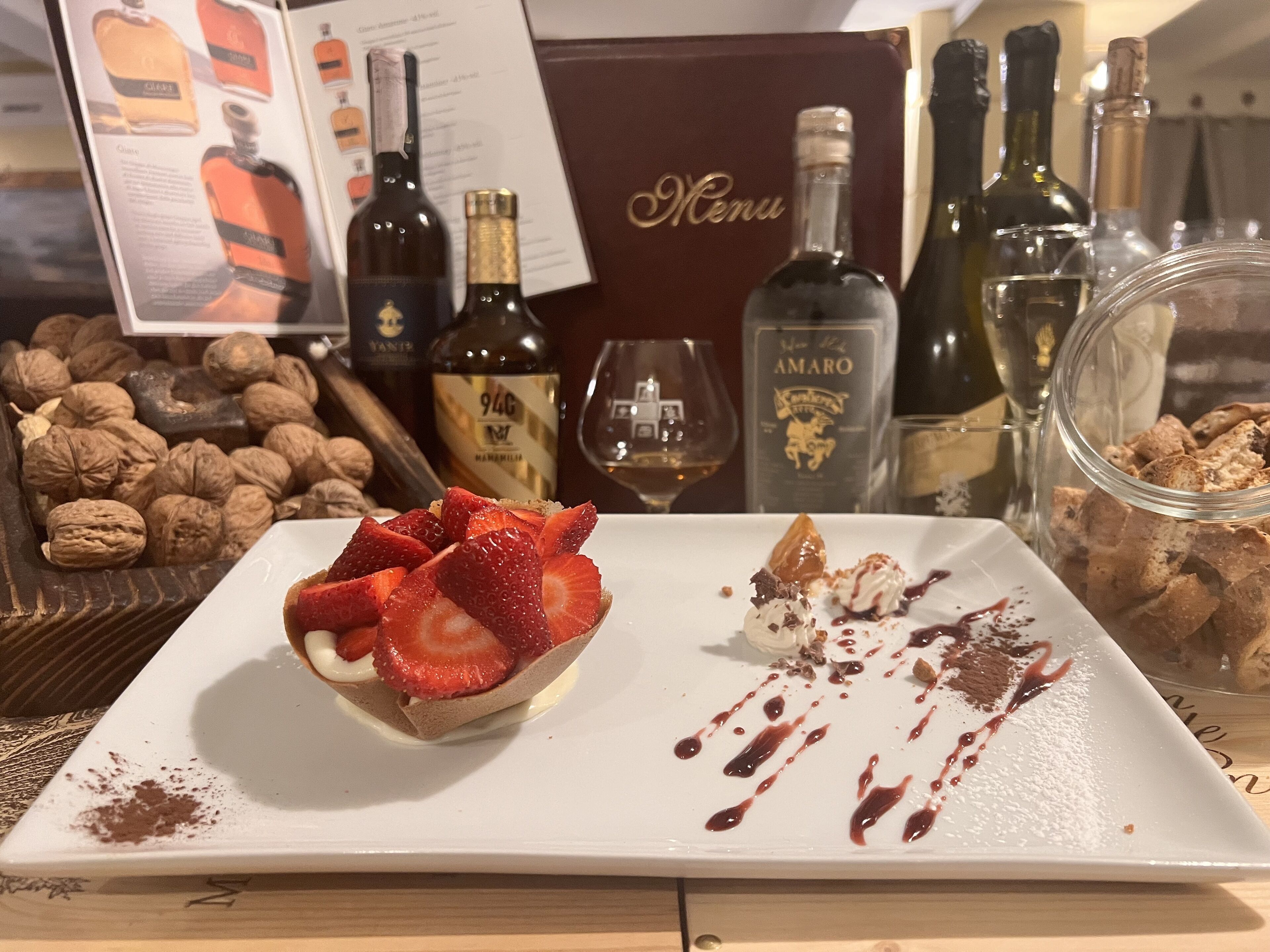 Un piatto dessert con fragole e cioccolato, circondato da bottiglie di vino e un menu.