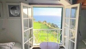 A room with a view of the ocean, a balcony with a metal railing, and a small table.
