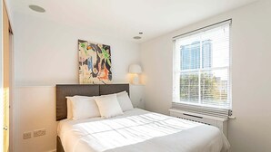 Ein Schlafzimmer mit einem großen Bett, einem Kopfteil, einem Fenster mit Jalousien, einer Wandlampe und einem Gemälde.