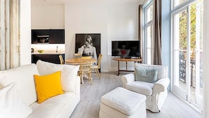 Ein modernes Wohnzimmer mit einem weißen Sofa, einem gelben Kissen, einem weißen Sessel und einem Fernseher. Es gibt eine Essecke mit Tisch und Stühlen, und im Hintergrund befindet sich eine Küche.
