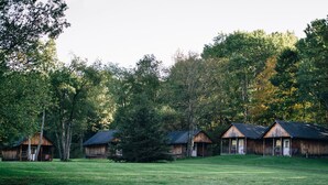 Une rangée de cabanes rustiques, avec un champ herbeux et une forêt dense en arrière-plan.