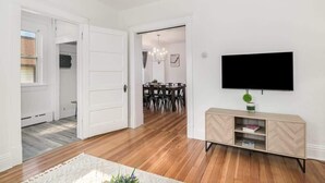 Una sala de estar moderna con piso de madera, una puerta blanca que da a un comedor y un televisor montado en la pared.