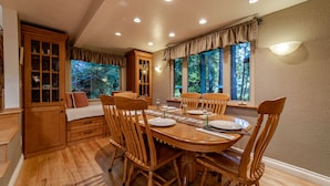 Comedor con mesa de madera dispuesta para seis personas, rodeada de sillas de madera. A la izquierda, un aparador de madera con puertas de cristal y, a la derecha, una ventana con cortinas.