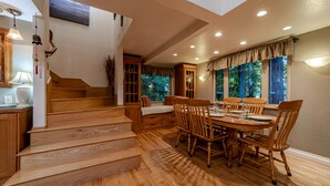 Un comedor con una mesa y sillas de madera, una escalera y vistas a árboles a través de amplios ventanales.