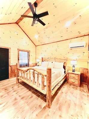 A wooden cabin bedroom with a bed, a nightstand, a ceiling fan, and an air conditioning unit.