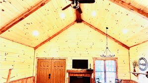 Een ruime kamer met een hoge plafond, houten wanden, een plafondventilator, een open haard, een bar met krukken en een aan de muur bevestigde televisie.