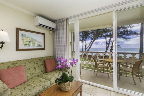 Ein Zimmer mit einer Couch, einem Tisch mit Pflanze, einem gerahmten Bild an der Wand und einem Balkon mit Stühlen und Meerblick.
