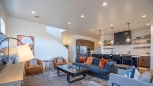 Un salon moderne avec un canapé, une table basse et des fauteuils, une cuisine avec un coin repas, et un escalier menant à un niveau supérieur.