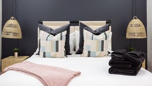 Ein Bett mit weißem Gestell, einer rosa Decke und einem Stapel schwarzer Handtücher. Zwei Hängelampen mit Flechtenschirmen sind vorhanden.
