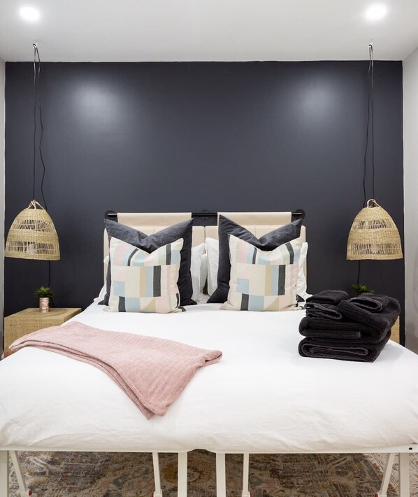 A bed with a white frame, a pink blanket, and a stack of black towels. There are two hanging lamps with woven shades.