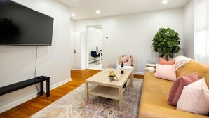 Ein modernes Wohnzimmer mit Flachbildfernseher, Couchtisch aus Holz, einer Couch mit Kissen, einem gemusterten Teppich und einer Zimmerpflanze.