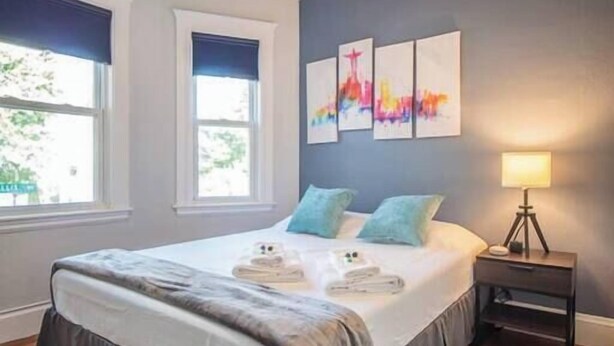 A bedroom with a bed, a ceiling fan, artwork on the wall, and a nightstand with a lamp. - Cozy House close to TUFTS, Harvard, MIT 3BR (Medford)