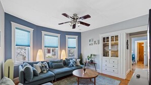 A modern living room with a blue sofa, a wooden coffee table, and a ceiling fan. - Cozy House close to TUFTS, Harvard, MIT 3BR (Medford)