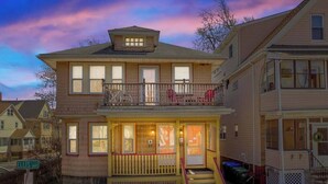 A two-story house with a balcony on the second floor, a front porch with a railing, and a visible front door. - Cozy House close to TUFTS, Harvard, MIT 3BR (Medford)
