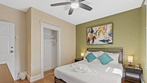 Un dormitorio ordenado con cama, mesitas de noche, ventilador de techo y un cuadro en la pared.