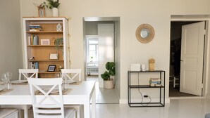 Ein Essbereich mit einem weißen Tisch und Stühlen, ein Bücherregal mit Büchern und Dekorationsobjekten, eine kleine Pflanze und ein wandmontierter Spiegel.