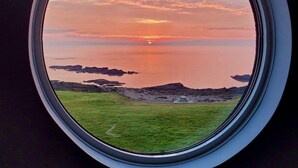 Et rundt vindue med udsigt til solnedgang over et roligt hav og en græsplæne.