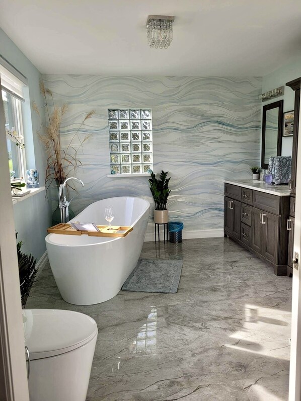 Un baño moderno con una bañera independiente, un tocador con armarios de madera oscura, un espejo grande y una pared decorativa con un patrón ondulado.