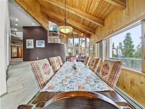 Une salle à manger en bois avec une longue table, des coussins à motifs et une vue sur un chalet, visible à travers de grandes fenêtres.