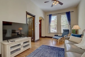 A living room with a flat-screen TV on a wooden cabinet, a blue armchair, a sofa, and a ceiling fan.