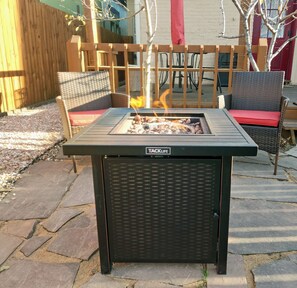 A black outdoor grill with a fire burning, surrounded by stone tiles and wicker furniture.