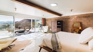 Une chambre moderne avec un grand lit, un banc en bois, un télescope et une vue sur les montagnes, visible à travers de grandes portes vitrées.