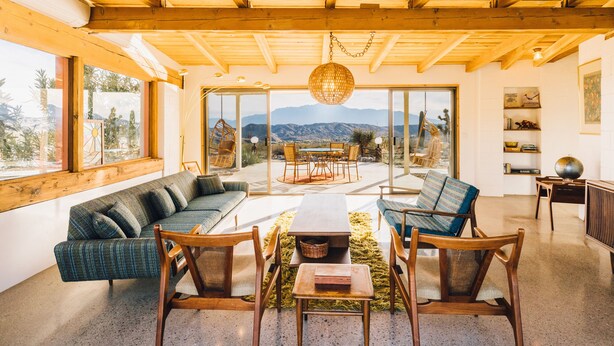Un vaste espace de vie doté d’un plafond en bois, de grandes fenêtres offrant une vue sur les montagnes, d’un coin salon central avec une table et d’un luminaire suspendu.