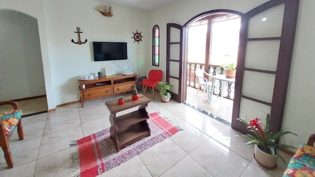 Una sala de estar con un mueble bajo de madera para el televisor, un sillón rojo, una mesa de centro de madera y una alfombra roja y blanca. Hay plantas en macetas y un ventanal con vistas al exterior.