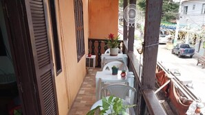 Un balcón estrecho con sillas y mesas de plástico blanco, plantas en macetas y vista a la calle.