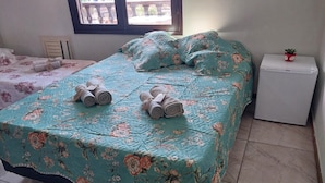 Un dormitorio con colcha estampada, dos toallas enrolladas sobre la cama, un pequeño armario blanco y una ventana con vistas al exterior.